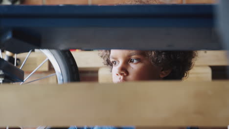 Close-up-of-pre-teen-mixed-race-boy-inspecting-his-racing-kart-in-the-garage,-seen-through-kart-frame,-selective-focus
