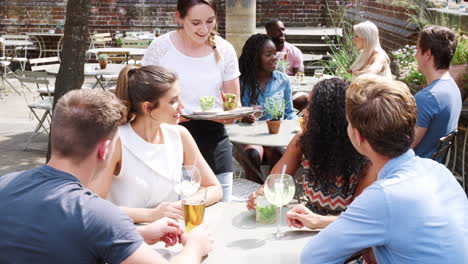 Group-Of-Friends-Meeting-For-Drinks-At-Outdoor-Tables-In-Restaurant-Being-Served-Snacks-By-Waitress