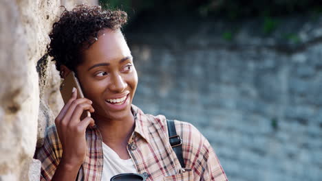 Millennial-black-woman-with-short-curly-hair-wearing-a-plaid-shirt-leaning-on-stone-wall-talking-on-smartphone-and-drinking-a-takeaway-coffee,-close-up