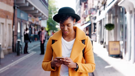 Millennial-black-woman-wearing-a-yellow-pea-coat-and-a-homburg-hat-using-her-smartphone-standing-on-a-street-and-walking-out-of-shot,-close-up