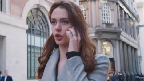 Close-up-of-smartly-dressed-millennial-businesswoman-walking-in-the-street-and-talking-on-her-smartphone,-focus-on-foreground