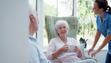 Senior-Couple-Sitting-In-Chair-And-Talking-With-Nurse-In-Retirement-Home