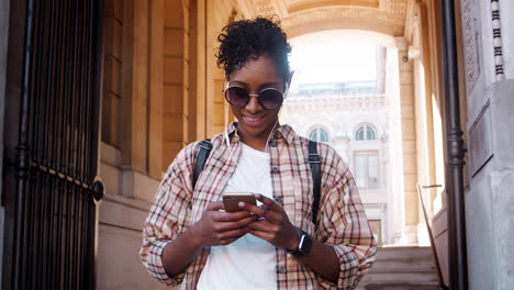 Close-up-of-fashionable-young-black-woman-wearing-sunglasses-and-plaid-shirt-using-her-smartphone-standing-outside-the-entrance-of-a-historical-building,-low-angle