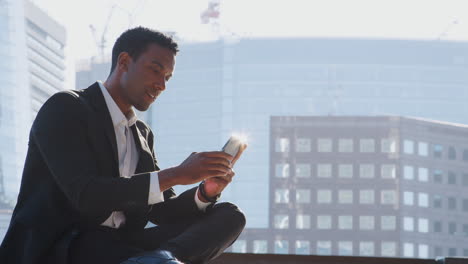Close-up-of-millennial-black-businessman-wearing-a-suit-and-a-white-shirt-sitting-on-the-River-Thames-embankment-using-his-smartphone,-close-up
