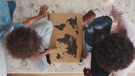 Close-up-overhead-view-of-a-mixed-race-pre-teen-boy-and-his-father-sitting-cross-legged-on-the-floor-opposite-each-other-playing-an-educational-game-with-a-world-map-and-map-pins