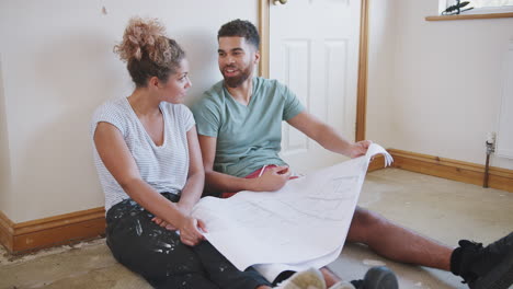 Couple-Sitting-On-Floor-Looking-At-Floor-Plans-In-Empty-Room-Of-New-Home