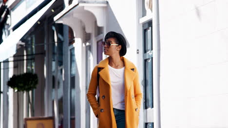 Fashionable-young-black-woman-wearing-blue-jeans-and-an-unbuttoned-yellow-pea-coat-walking-in-the-street-past-shops-on-a-sunny-day,-smiling,-close-up