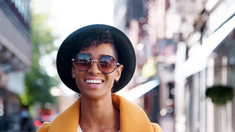 Millennial-black-woman-wearing-a-yellow-pea-coat,-sunglasses-and-a-homburg-hat,-standing-on-a-city-street-smiling-to-camera,-close-up