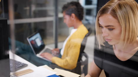 Two-young-women-working-together-on-computers-in-a-glass-office-cubicle,-one-turning-around-to-the-other,-rack-focus,-seen-through-glass