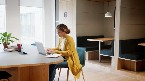 Young-adult-woman-working-at-laptop-computer-sitting-in-an-office-while-her-colleague-enters-the-room,-handheld