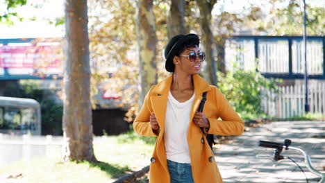 Fashionable-young-black-woman-wearing-a-hat,-sunglasses,-blue-jeans-and-a-yellow-pea-coat-walking-along-a-treelined-city-street-on-a-sunny-day,-looking-around,-smiling,-front-view