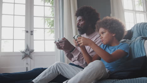 Black-middle-aged-father-and-his-pre-teen-son-sitting-on-the-floor-in-their-living-room-eating-and-playing-video-games-together,-close-up