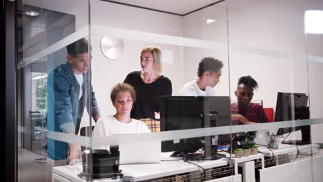Creative-business-team-gathered-around-a-laptop-computer-in-a-small-office-cubicle,-seen-through-glass