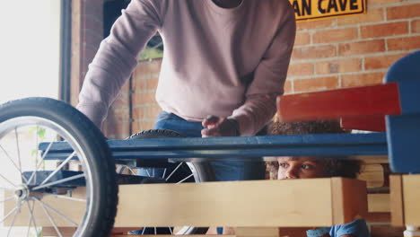 Close-up-of-middle-aged-black-father-and-his-pre-teen-son-standing-at-a-workbench-in-their-garage-building-a-racing-kart-together,-low-angle