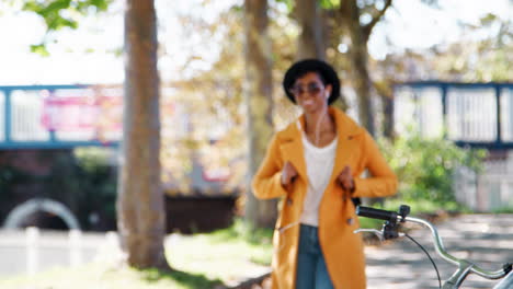 Fashionable-young-black-woman-wearing-a-hat,-sunglasses,-blue-jeans-and-a-yellow-pea-coat-walking-along-a-treelined-street-on-a-sunny-day-listening-music-with-earphones,-rack-focus