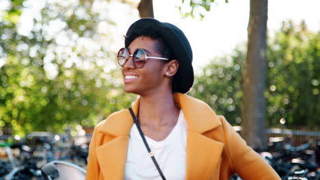 Fashionable-young-black-woman-standing-outdoors-wearing-sunglasses,-a-yellow-coat-and-a-black-hat-looking-to-camera-and-laughing,-close-up,-focus-on-foreground