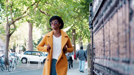 Millennial-black-woman-wearing-an-unbuttoned-yellow-coat-and-a-hat-walking-down-a-city-street-past-railings,-selective-focus