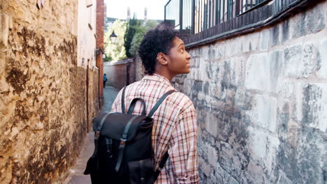 Young-black-woman-with-a-backpack-walking-in-a-narrow-alleyway-between-stone-walls,-back-view,-follow-shot