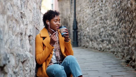 Young-woman-wearing-a-yellow-pea-coat-and-blue-jeans-sitting-in-an-alleyway-in-a-historical-city-talking-on-her-smartphone-using-earphones,-close-up