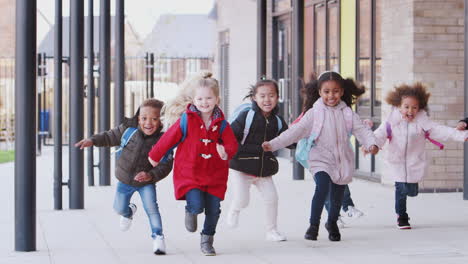 Happy-young-school-kids-wearing-coats-and-bags-running-in-a-walkway-outside-their-school-building