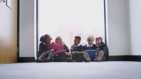 Five-primary-school-kids-sitting-on-the-floor-in-a-school-corridor-using-tablet-computers,-low-angle