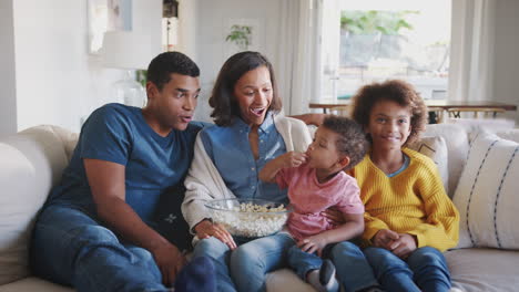 Junge-Familie-Sitzt-Zusammen-Auf-Dem-Sofa-In-Ihrem-Wohnzimmer,-Schaut-Fern-Und-Isst-Popcorn