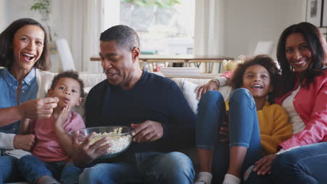 Three-generation-African-American-family-sitting-on-the-sofa-watching-TV-and-eating-popcorn,-panning-shot