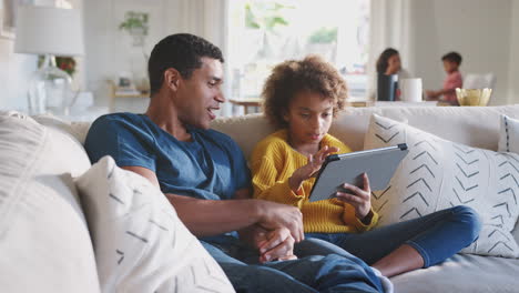 Father-and-his-pre-teen-daughter-on-sofa-using-tablet-computer,-mum-and-toddler-in-the-background