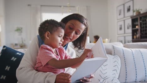 African-American-mum-sitting-on-sofa-in-living-room-reading-to-her-toddler-son,-on-her-knee,-close-up