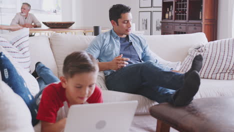 Dad-comes-to-sit-with-pre-teen-son-using-laptop-in-sitting-room,-mum-and-grandad-in-background