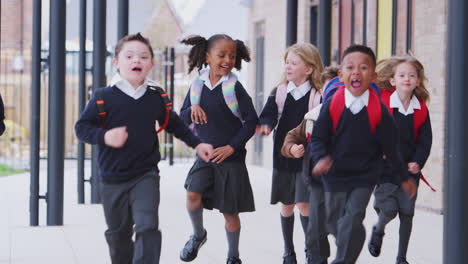 Happy-primary-school-kids-in-uniforms-running-on-a-walkway-outside-their-school-building,-front-view