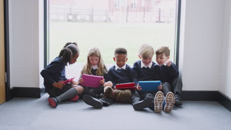Five-primary-school-kids-sitting-on-the-floor-in-a-school-corridor-with-tablet-computers