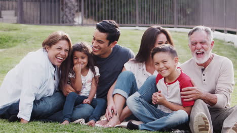 Three-generation-family-sitting-on-grass-in-the-park,-tickling-each-other-and-smiling-to-camera