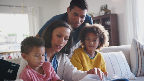 Young-African-American-mum-reading-a-book-to-her-two-children,-dad-looking-over-their-shoulders,-close-up