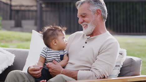 Senior-Hispanic-man-sitting-on-a-seat-in-the-garden-with-his-baby-grandson-on-his-knee,-close-up