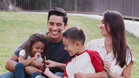 Glückliche-Junge-Hispanische-Familie,-Die-Auf-Dem-Gras-Im-Park-Sitzt,-Sich-Gegenseitig-Kitzelt-Und-In-Die-Kamera-Lächelt