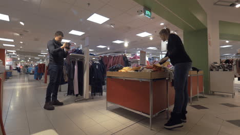 Man-with-mobile-shooting-woman-choosing-kids-clothing-in-the-store