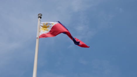 Bandera-Nacional-Filipina-Mientras-La-Cámara-Se-Inclina-Hacia-Arriba-Y-Se-Aleja-Un-Poco,-Revelando-La-Bandera-Ondeando-Hacia-La-Derecha-Y-El-Fantástico-Cielo-Azul-Y-Algunas-Nubes-Finas-Y-Esponjosas