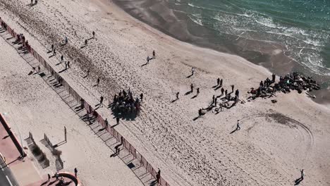 Rotating-aerial-view:-People-on-horses-moving-along-the-Palavas-beach-in-Southern-France,-surrounded-by-tourists