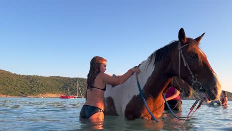 Funny-little-girl-in-swimsuit-attempts-to-get-on-horse-in-sea-water-in-summer-season