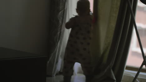 A-baby-girl-in-an-overall-playing-with-curtains-near-the-large-window