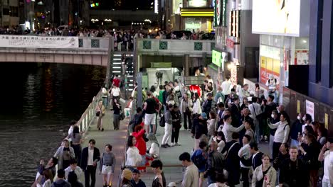 Busy-Crowds-Illuminated-By-Neon-Billboard-Light-Taking-Photos-Of-Glico-Man-Beside-Dotonbori-Canal-At-Night