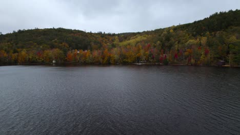 Flying-over-lake-near-colorful-fall-forest-on-hill-during-overcast-day