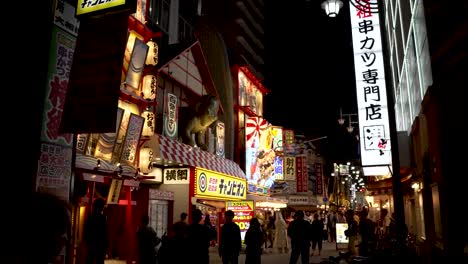 Silhouette-Of-People-Along-Street-In-Shinsekai-Area-Illuminated-By-Neon-Signs-At-Night