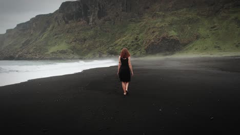 Hermosa-Pelirroja-Vestida-De-Negro-Caminando-Por-La-Playa-De-Arena-Negra,-Islandia,-Tiro-De-Seguimiento