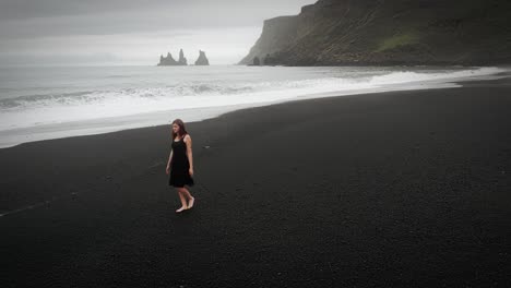 Young-beautiful-woman-in-black-dress-walking-on-black-sand-beach-Iceland,-epic-aerial-ocean-landscape