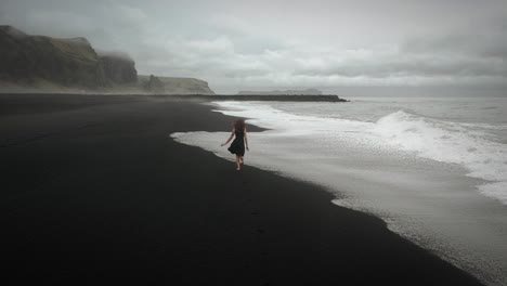 Aerial-young-beautiful-woman-in-black-dress-running-on-black-sand-beach-Iceland,-tracking-shot