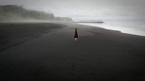 Young-beautiful-woman-in-black-dress-walking-on-black-sand-beach-Iceland,-foggy-dramatic-mountain-landscape,-tracking-shot