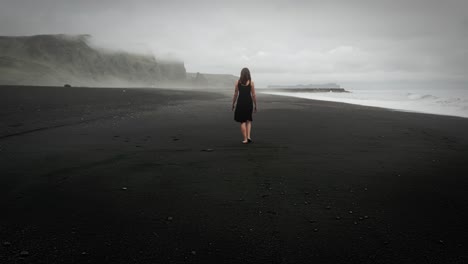 Young-beautiful-woman-in-black-dress-walking-on-black-sand-beach-Iceland,-misty-dramatic-mountain-landscape,-tracking-shot
