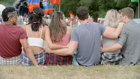 Friends-sit-on-grass-talking-at-a-music-festival,-back-view